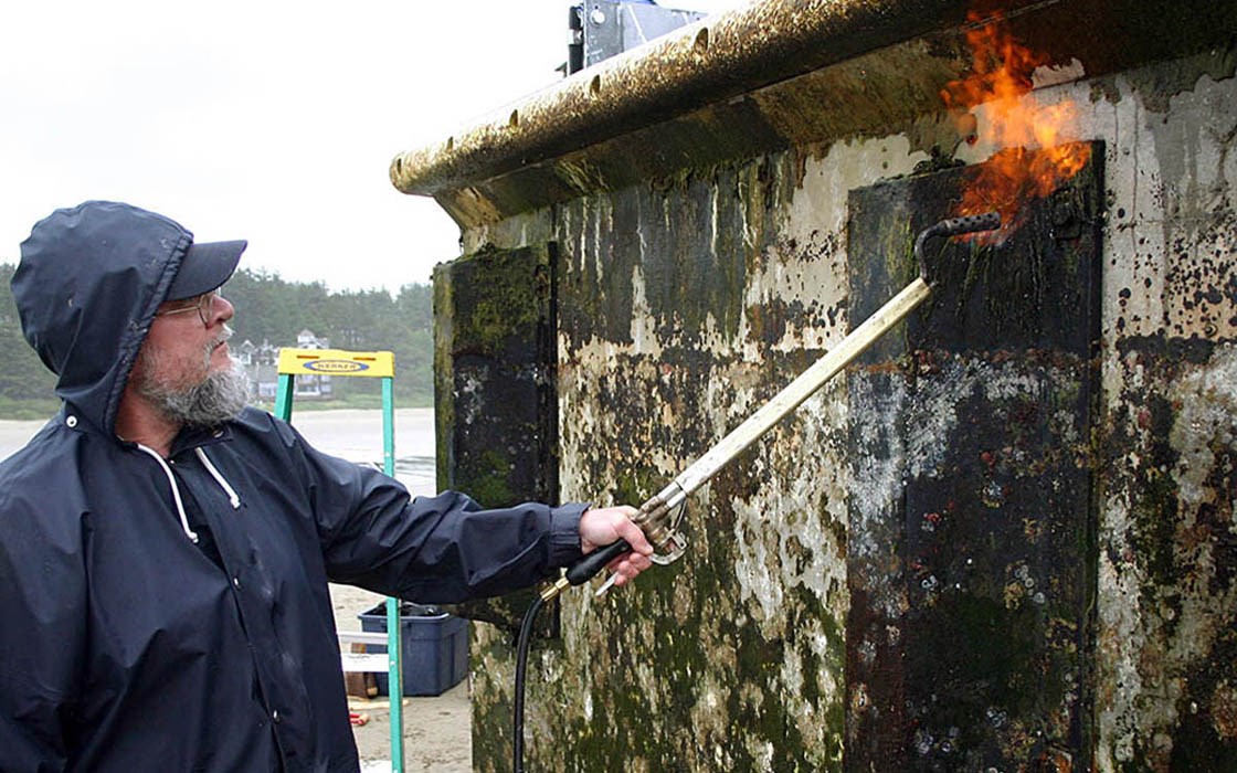 Worker cleaning the stranded dock The structure was scraped clean then, low-pressure torches were used to sterilize the dock. All removed material was bagged, hauled up the beach well above the high tide line and stored temporarily. Agate Beach, Oregon, USA. June 2012.