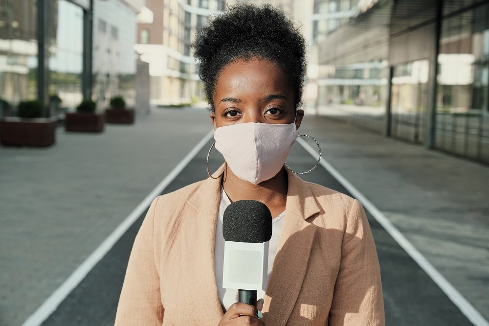Portrait of African young reporter in protective mask holding microphone and looking at camera while standing in the city