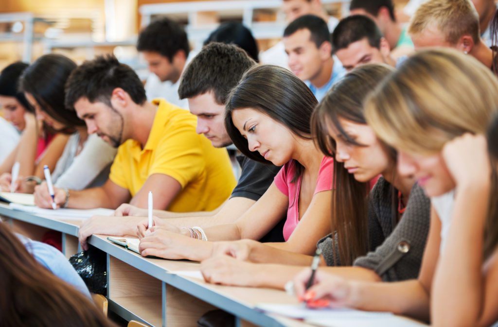 Large group of students writing. "Group of college students in the university amphitheatre, they are sitting and doing an exam. The focus is on the brunette in pink t-shirt."